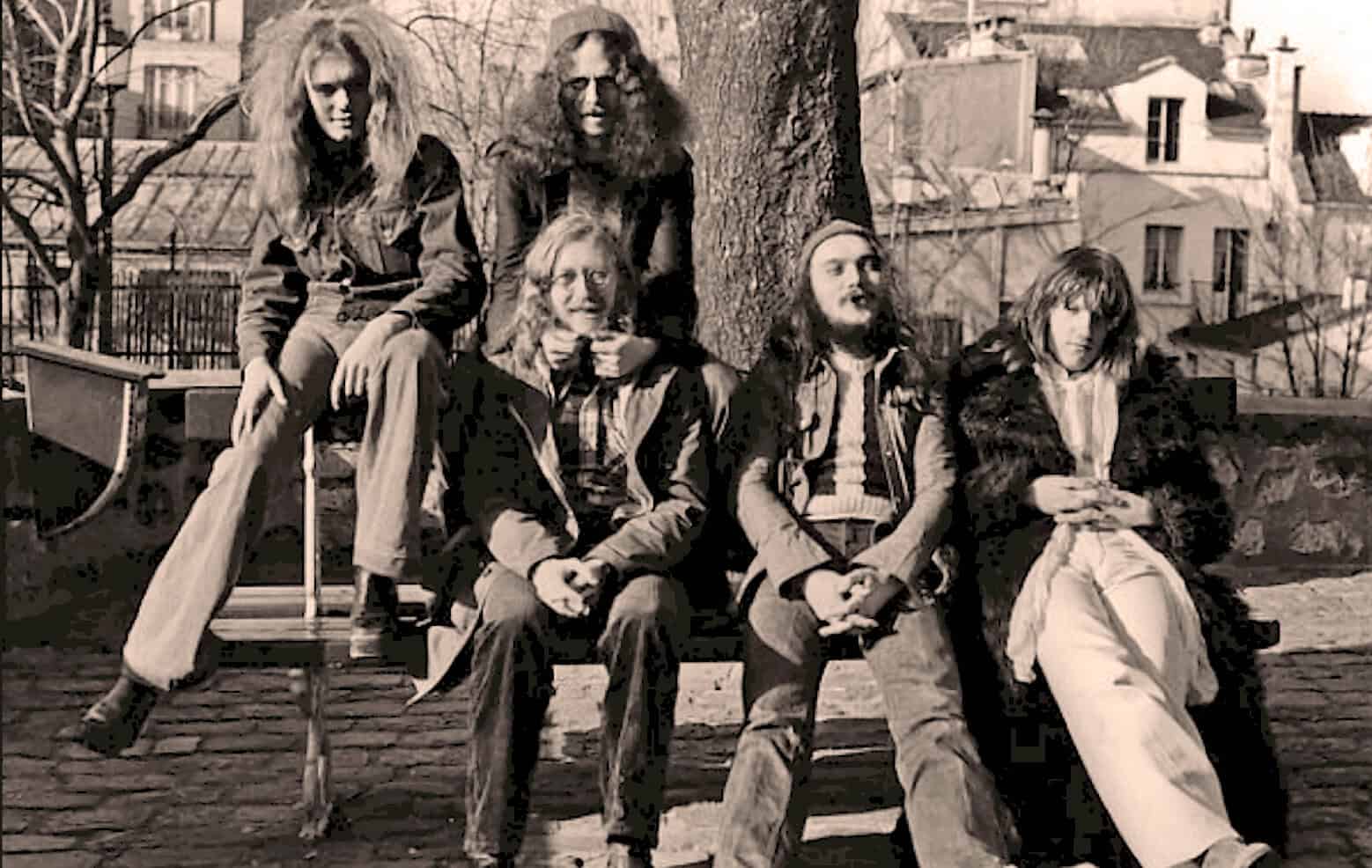 Wild Irish Roses band members sitting on a park bench under a large tree in an urban setting, black and white photo, casual clothing, music enthusiasts, alternative culture, 1970s music scene, nostalgic music archive, outdoor photo.