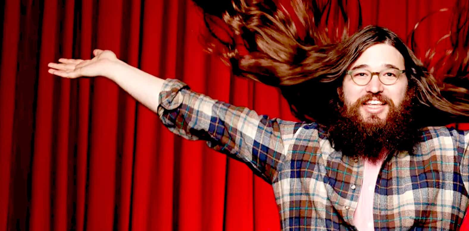 Vibrant photo of a cheerful man with long hair and beard, wearing glasses and a plaid shirt, posing in front of red curtains, emphasizing music, entertainment, and creative arts themes for SEO.