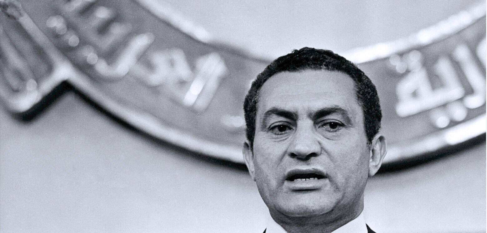 Historical black and white photo of a man speaking at a press conference, with a microphone and a news logo in the background, representing archives of news, history, and music from Past Daily.