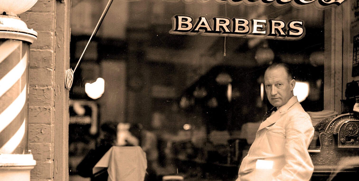 Vintage barber shop scene with a barber in a white coat standing inside the window, classic "Barbers" sign above, nostalgic atmosphere, sepia tone, highlighting traditional grooming services and historic interior.