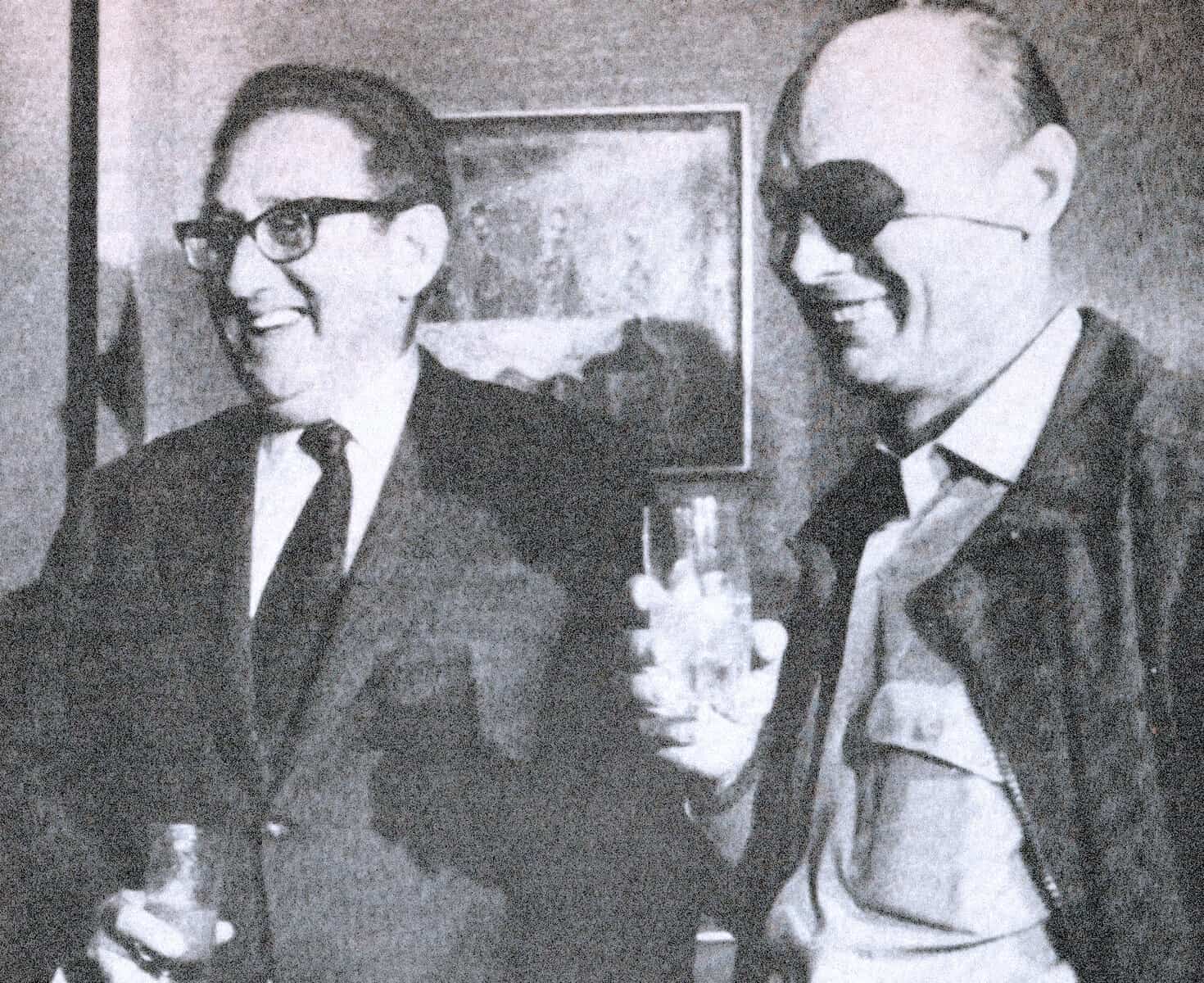 Close-up of two men wearing glasses and suits, smiling and enjoying drinks during a social event, with a framed picture on the wall behind them.