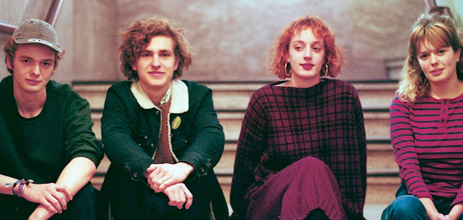 A young band of four individuals sitting on staircase with vintage clothing and hairstyles, representing music and youth culture from the late 20th century.
