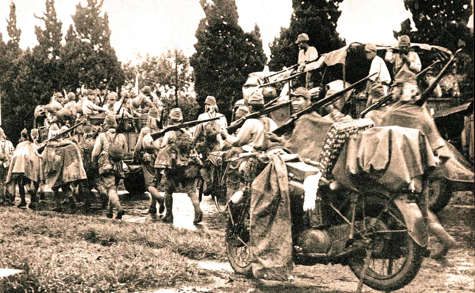 Historical photograph of early 20th-century soldiers and civilians gathered around military vehicles and horses, capturing a moment of wartime mobilization and historical significance.