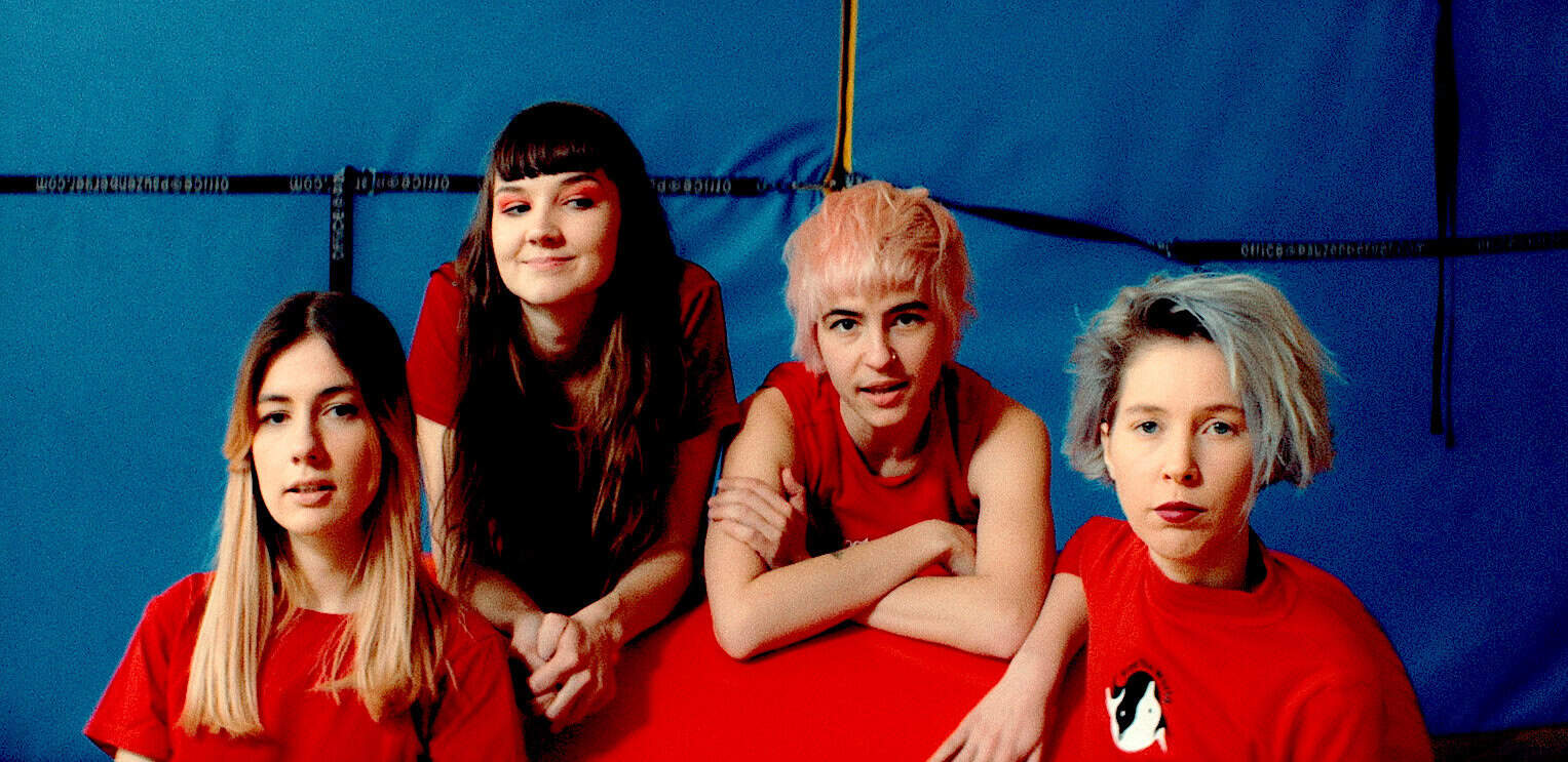 Girls band performing, alternative music, young women with bold hairstyles and red shirts at a concert, energetic female musicians in a vibrant stage setting, diverse female pop artists playing in a music event, relevance: high for music history, band performances, and female artists on Past Daily sound archive.