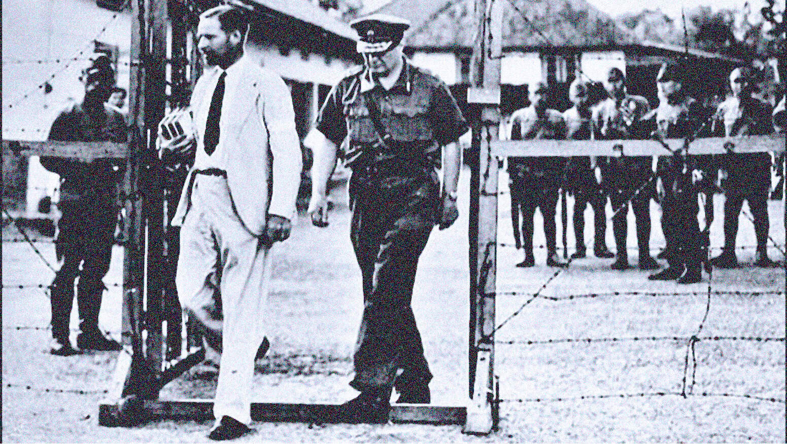 Man walking through a gated entrance with soldiers and civilians in the background, historical black-and-white photo highlighting era-specific news, history, and social context.