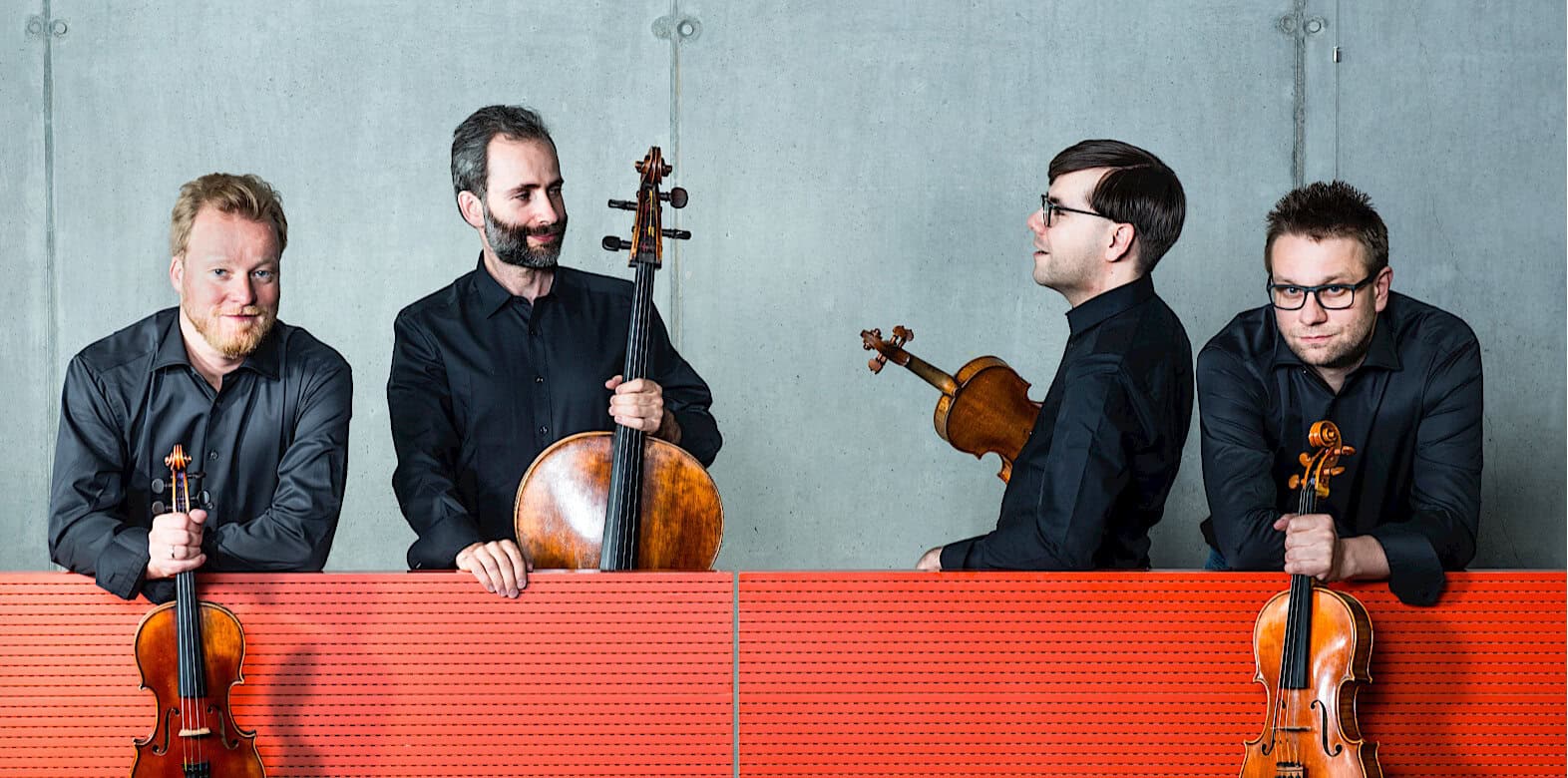 Elegant classical musicians with violins and a cello, dressed in black, standing against a plain wall, emphasizing the artistry and history of orchestral music.