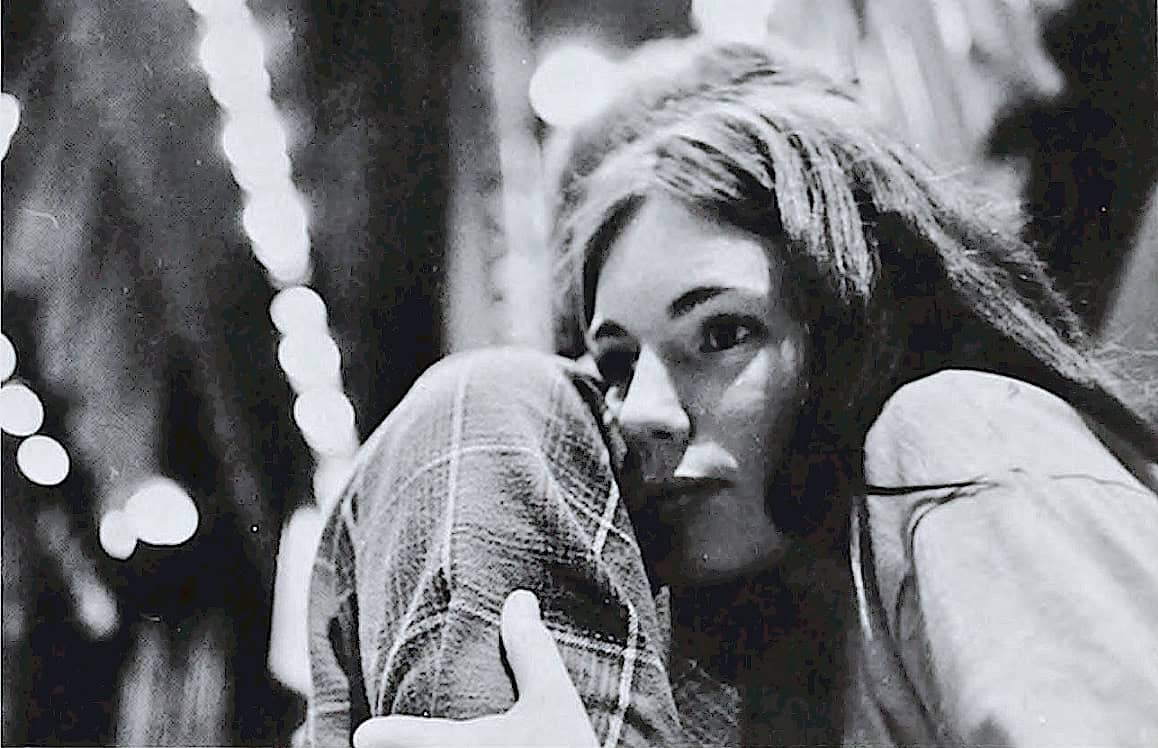 A black and white photo of a woman with long hair, gazing thoughtfully into the distance, holding a hat or piece of clothing close to her face, with blurred decorative lights in the background.