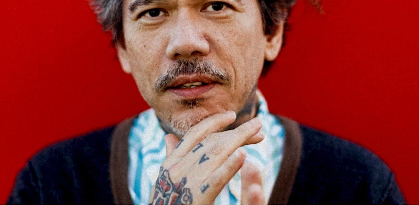 Portrait of a mature man with tattoos on hand, natural light, red background, expressive face, music and news archive theme, vintage style, podcast content, historical audio recordings, contemporary media, past daily archive, sound archive of news and music, historical sound recordings.