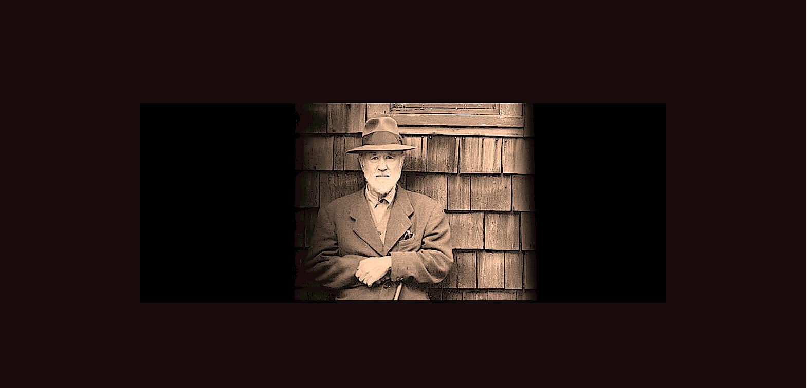 Old man with a fedora hat and suit standing against wooden shingles, representing history and vintage style.
