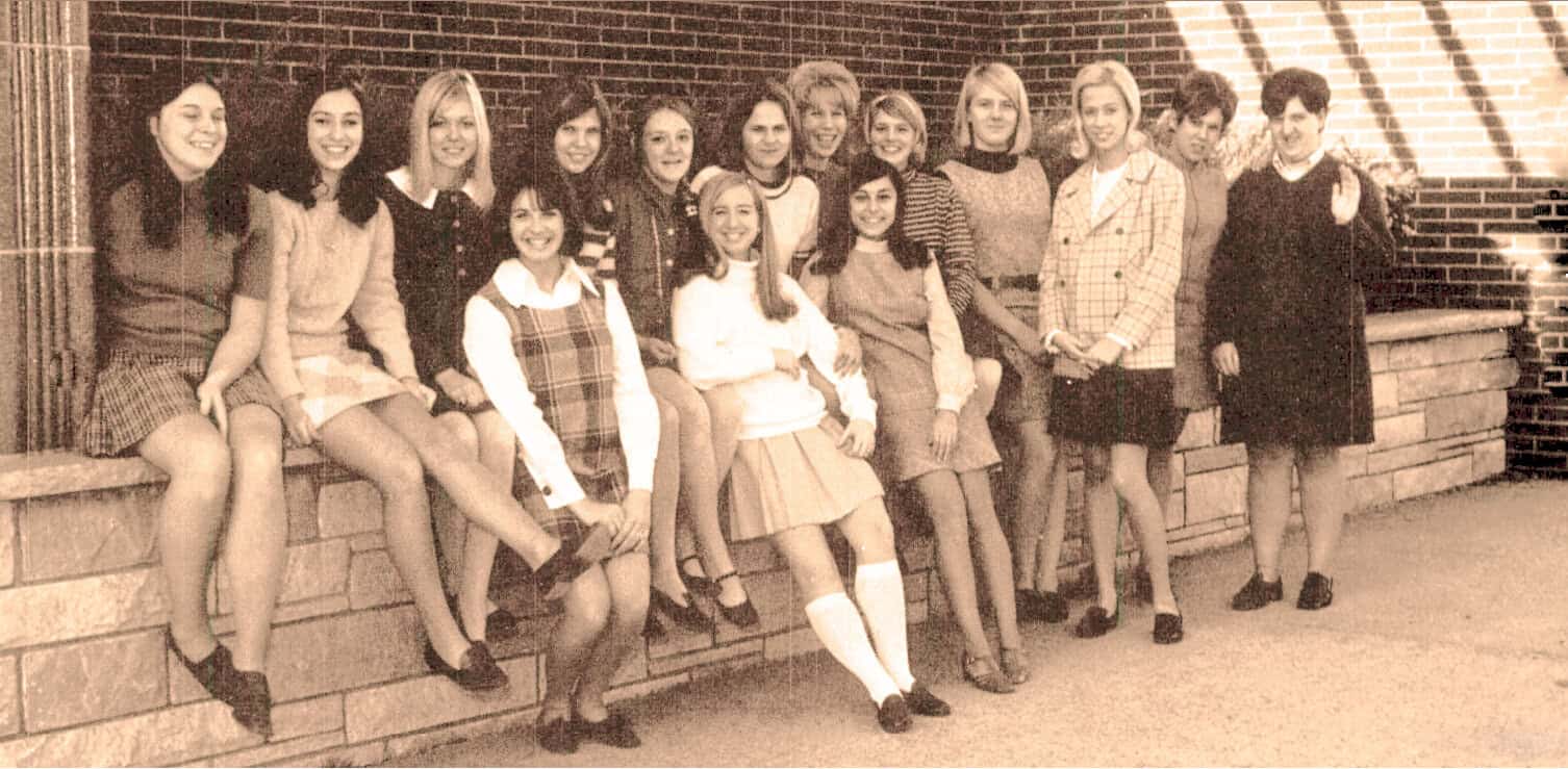 Vintage black and white photo of high school girls in school uniforms, smiling and posing together outside a brick building, capturing a nostalgic moment in education and youth history.