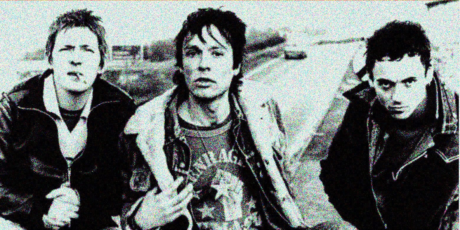 Black-and-white photo of three young men dressed in punk style, posing outdoors near a body of water, capturing a vintage music and news archive vibe.