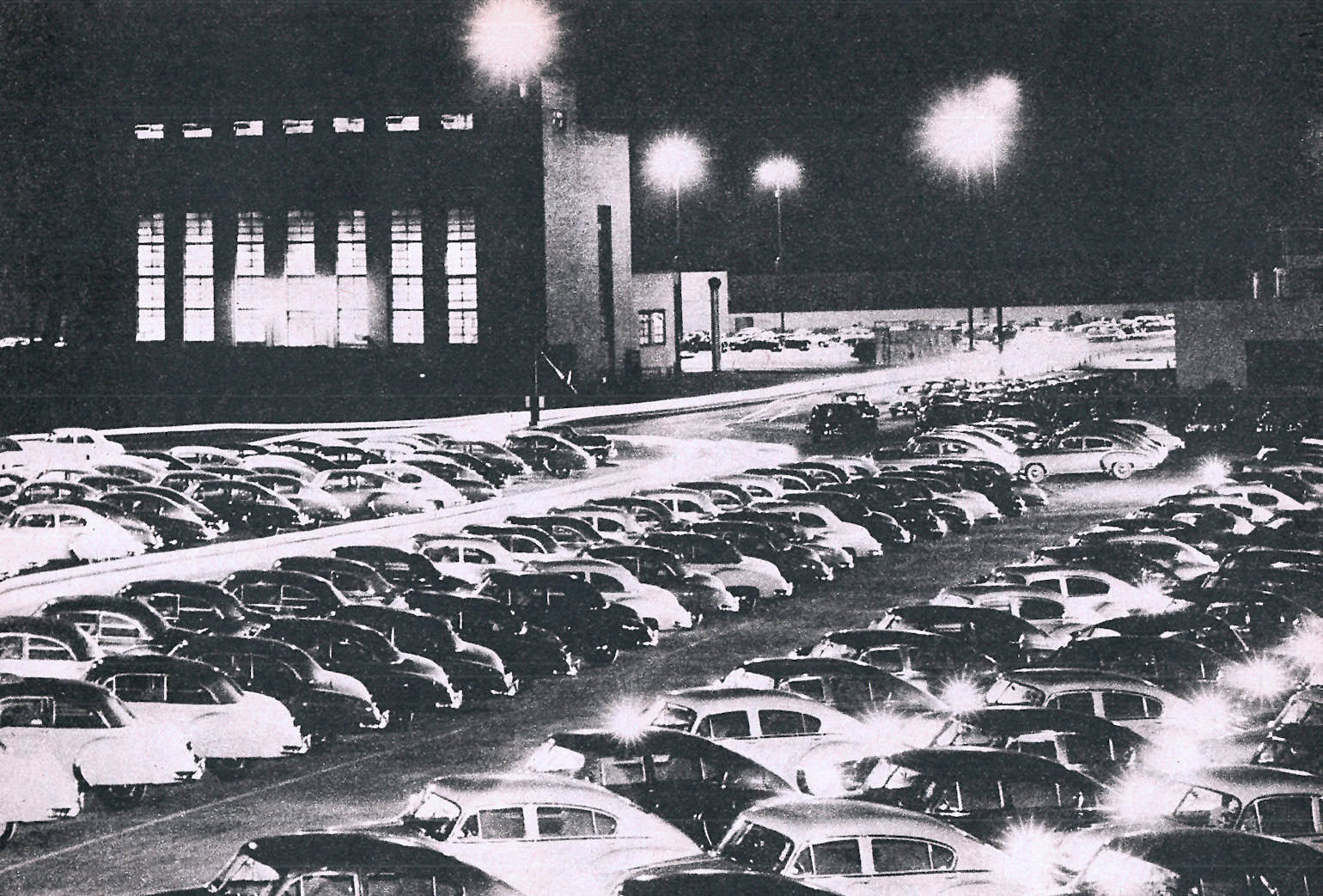 Packed parking lot at night with vintage cars under bright streetlights, capturing a nostalgic scene of mid-20th-century urban life, perfect for historical or music archive websites.