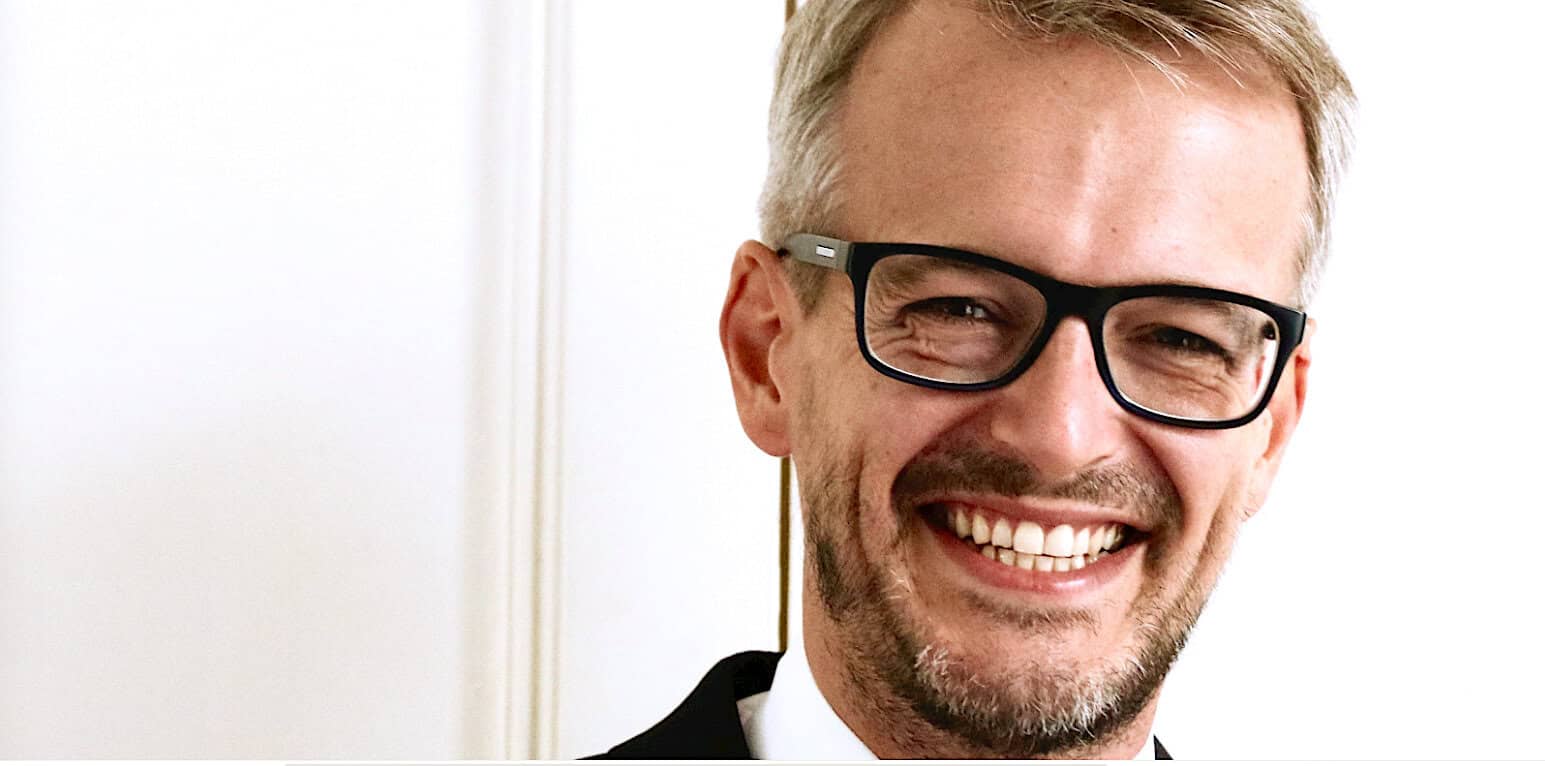 Professional man smiling, wearing glasses, indoor setting, happy expression, business attire, corporate environment, friendly colleague, positive mood, modern office, leadership, smiling executive.