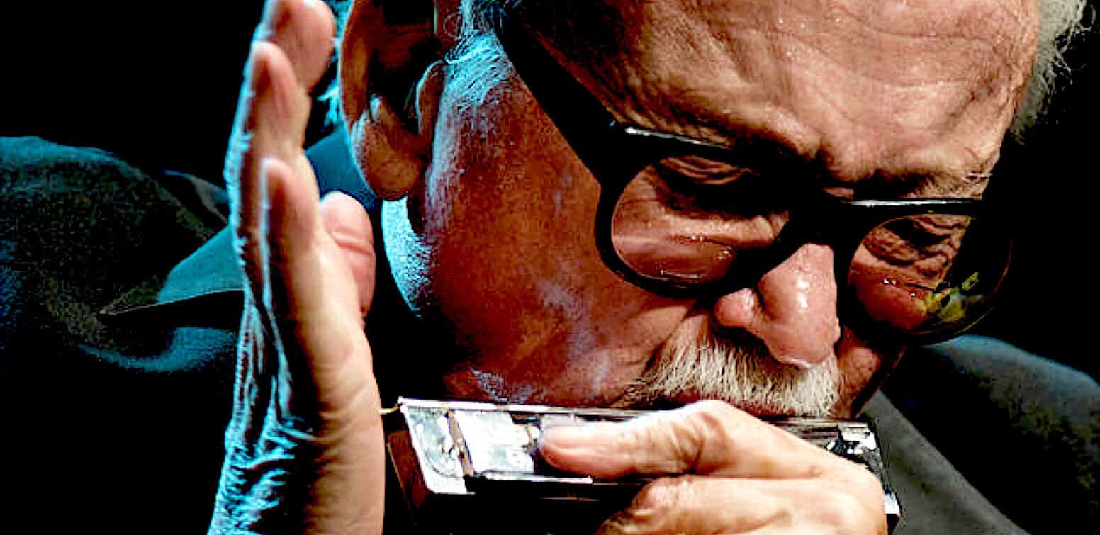 Portrait of an elderly man with glasses playing harmonica, capturing a moment of musical nostalgia and history at Past Daily sound archive.