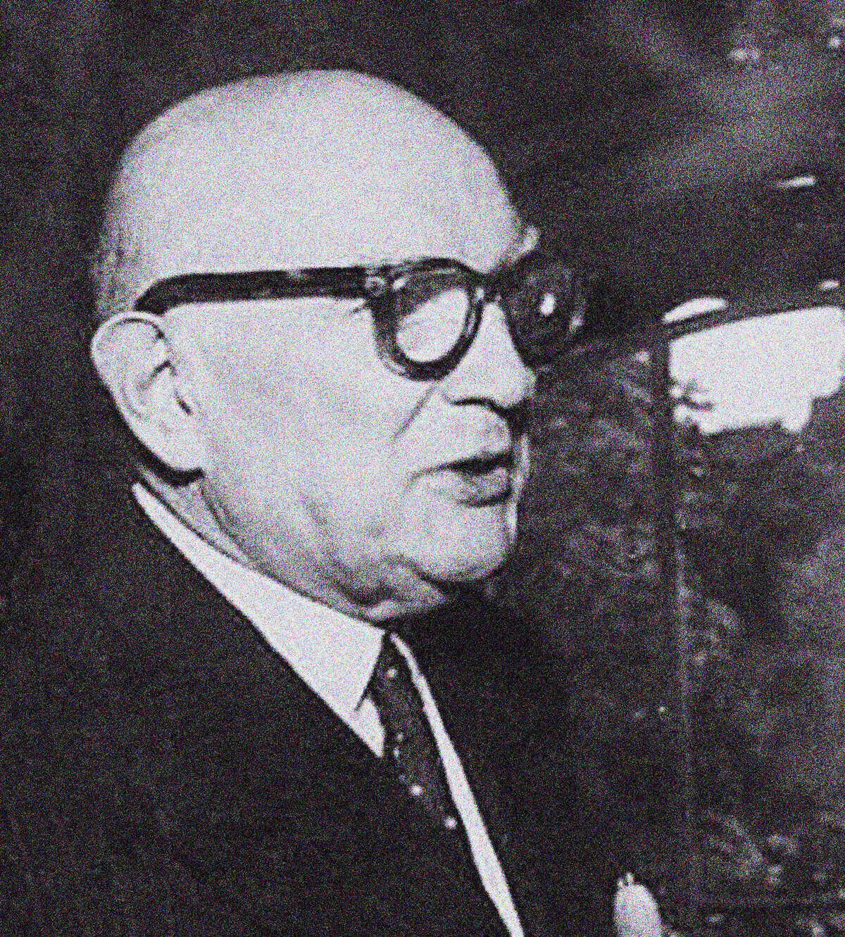 Bald man with glasses speaking during a broadcast, historical news and music archive.