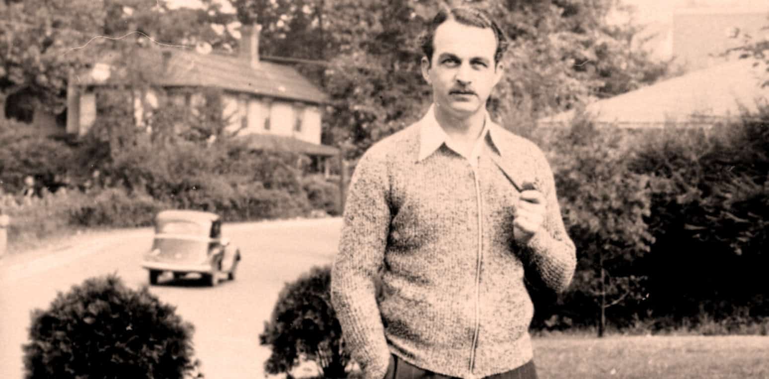 A vintage black and white photo of a man in a sweater holding a pipe, standing outdoors in a suburban neighborhood with trees and houses, capturing a historic moment related to music, news, and history archives.