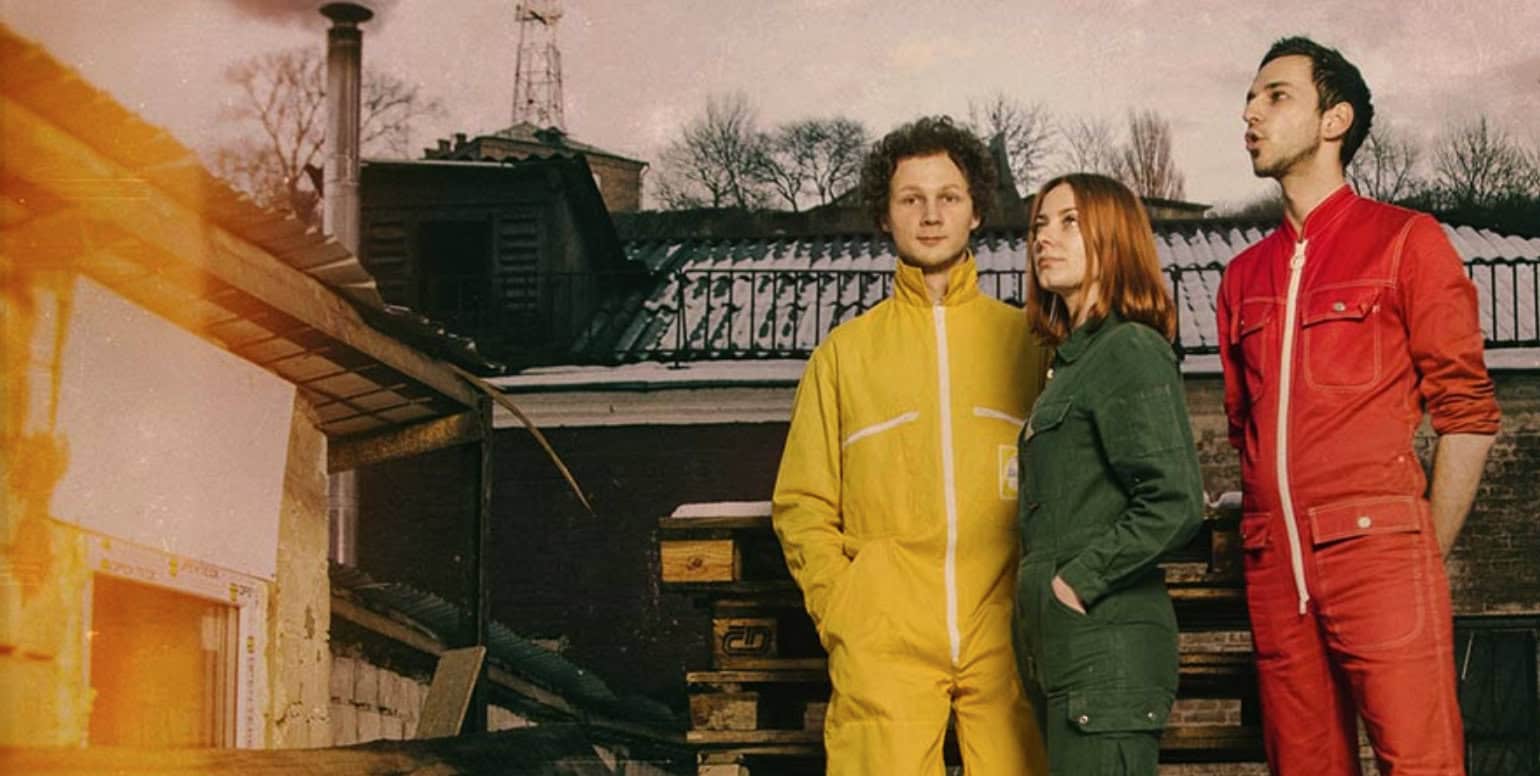 Colorful jackets of three young adults standing outdoors near a construction site with a background of rooftops and trees, representing modern urban fashion and youth culture.