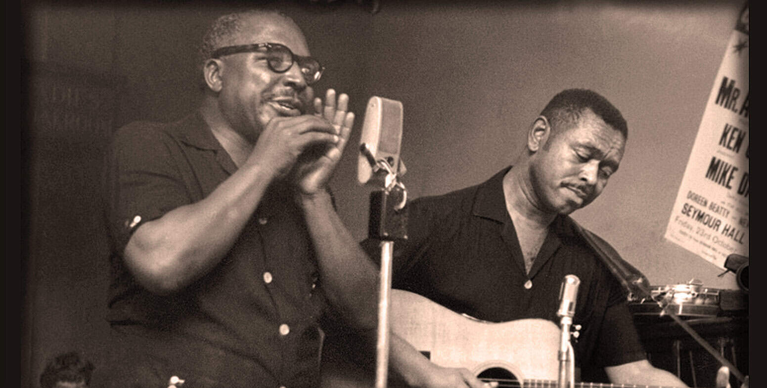 Vintage black and white photo of two male musicians recording a music session, one singing into a microphone and the other playing guitar, showcasing historic music and news archive from Past Daily.
