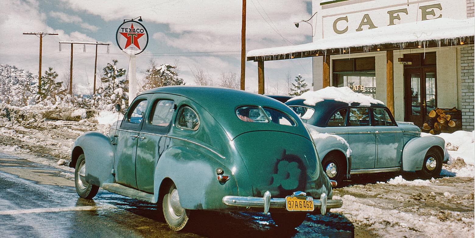 Vintage blue cars parked outside a cafe in a snowy winter scene, with a Texaco sign in the background, showcasing classic automobile entertainment and historic Winter ambiance.