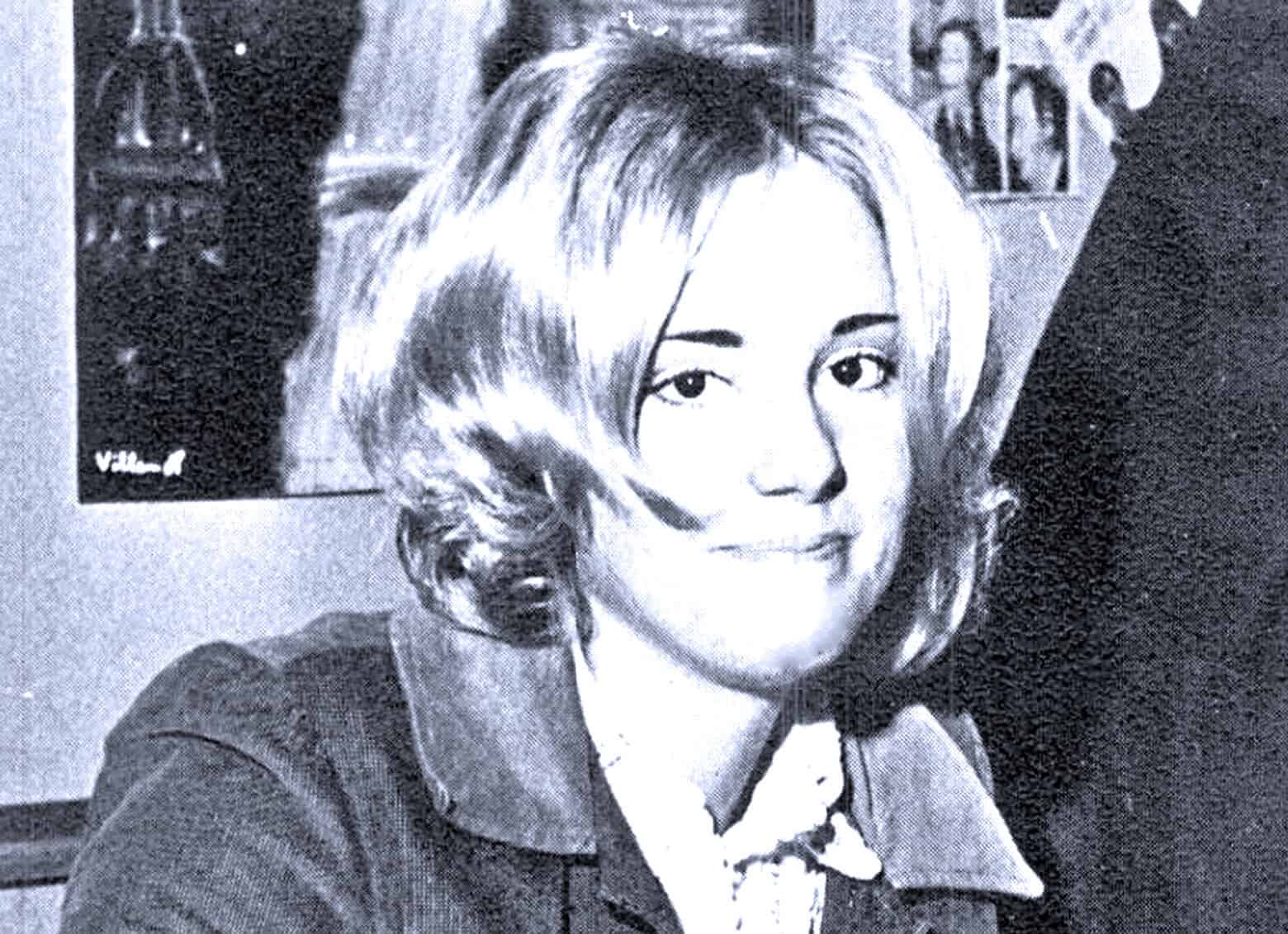 Vintage black and white photo of a woman with short, wavy hair smiling, seated indoors with wall art in the background, highlighting nostalgic music and news archive themes.