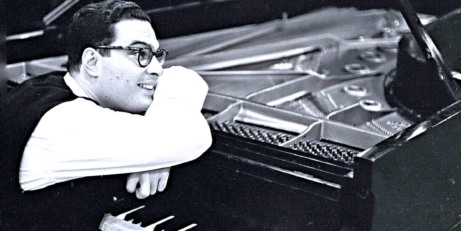 Soft focus black and white photo of a young man with glasses leaning on a grand piano, smiling, capturing a moment of musical creativity and jazz influence, showcasing vintage music history and cultural heritage.