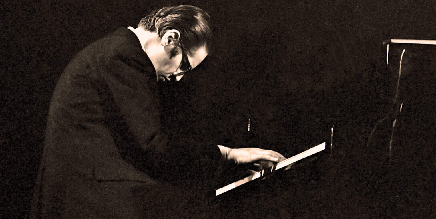 A black and white photo of a jazz pianist wearing glasses, playing a grand piano in a dimly lit setting.