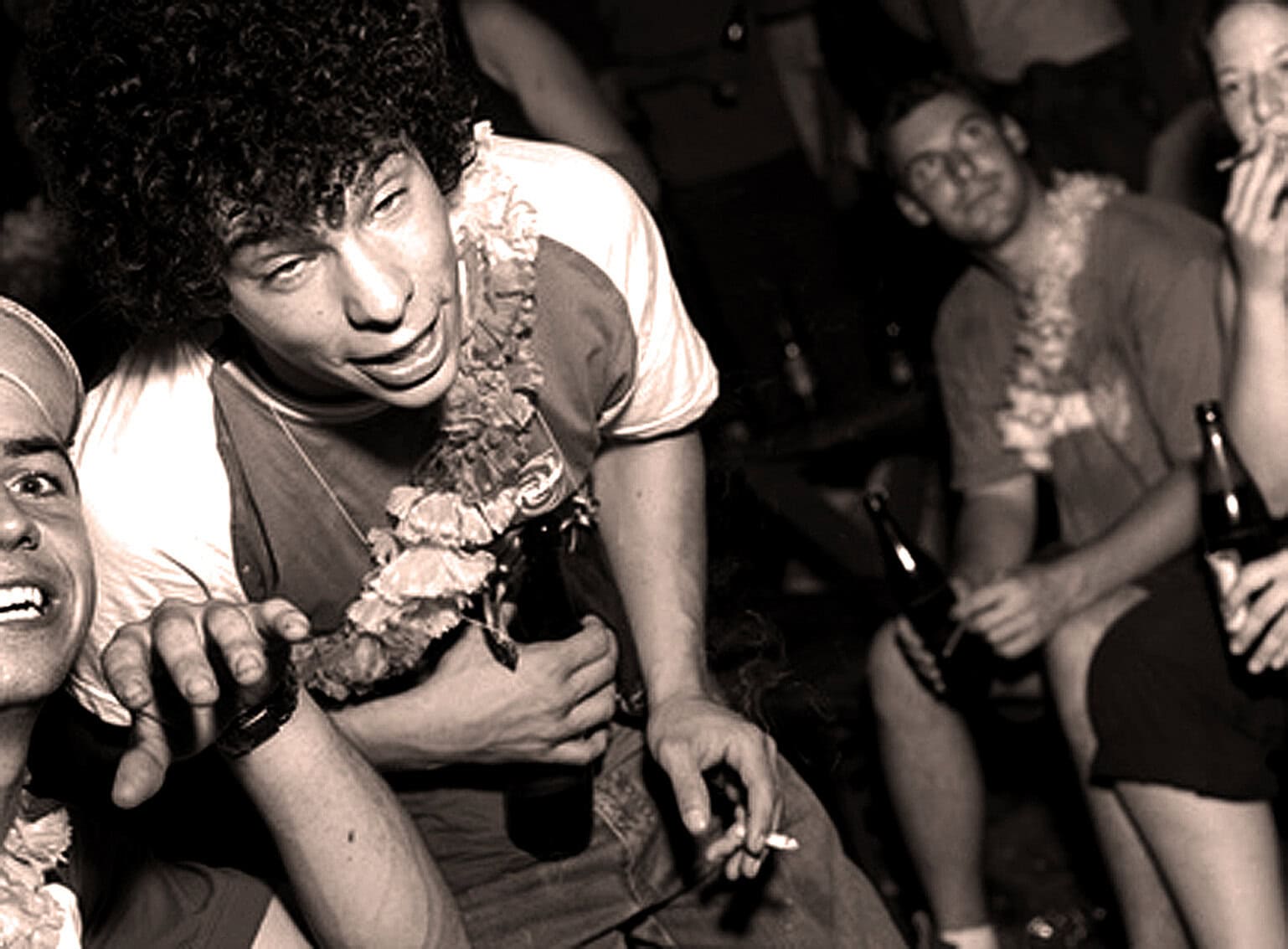 A young man with curly hair enjoying a lively party scene, surrounded by friends holding drinks, capturing a moment of music, fun, and social history on a vibrant night out.