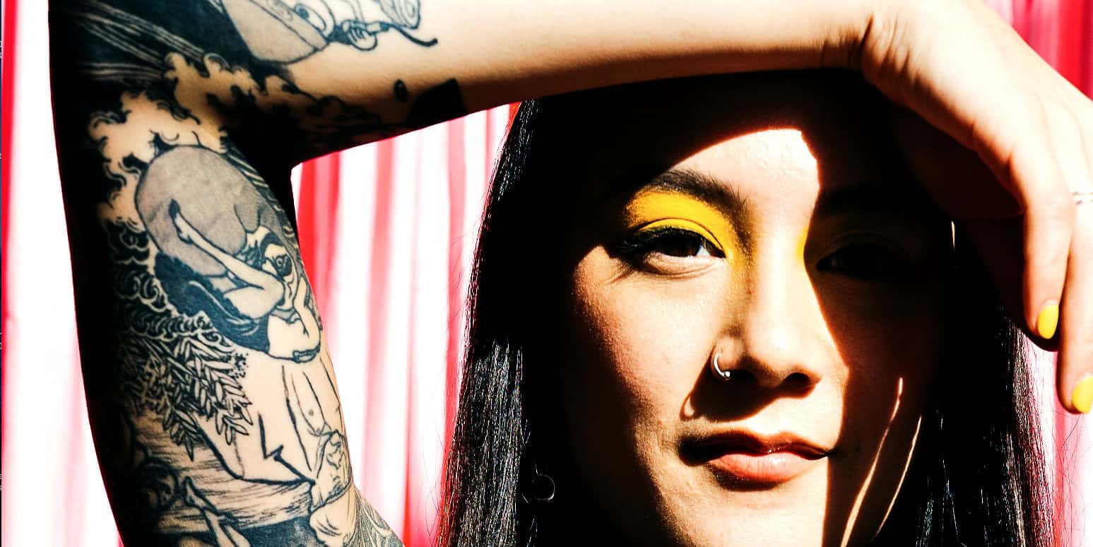 Bright woman with bold yellow eye makeup and tattoos, posing with hand over head in front of red striped background, emphasizing vibrant colors and tattoo art.
