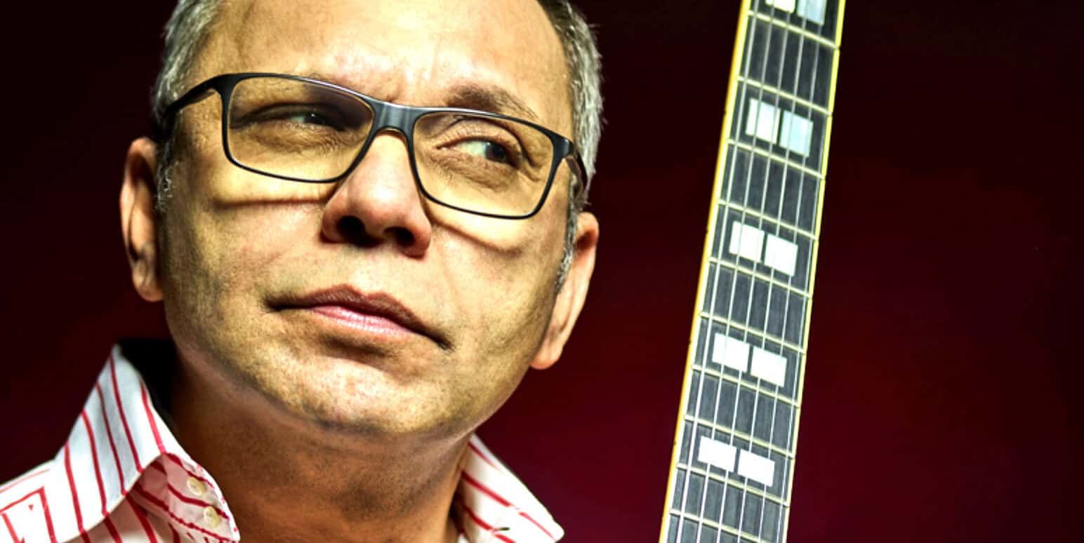 Close-up of a middle-aged man wearing glasses, holding an electric guitar, representing music history and legendary artists.