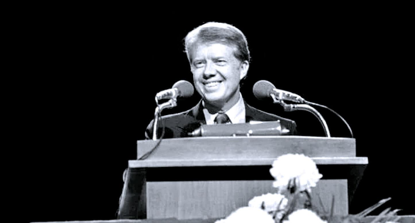Historical black and white photo of a man speaking at a podium with microphones, representing news, history, and music archive content from Past Daily.