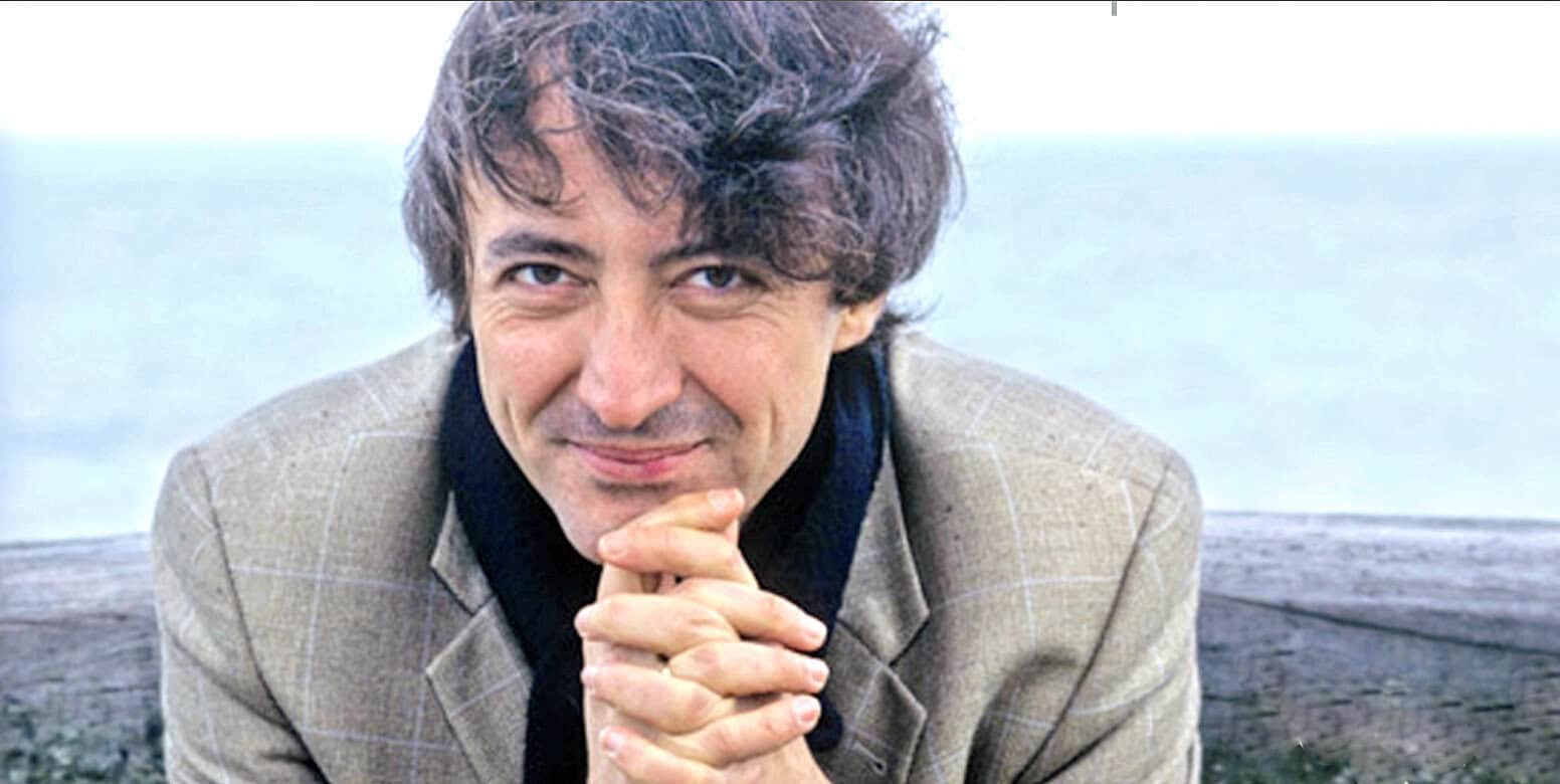 Vintage portrait of a man with dark curly hair, smiling thoughtfully outdoors near water, representing historical music and news archive theme of Past Daily.