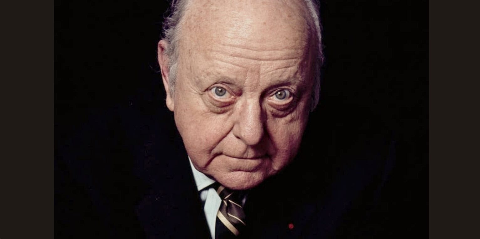 Close-up portrait of an elderly man with gray hair and blue eyes, wearing a dark suit and striped tie, symbolizing historical news coverage and timeless music archives.