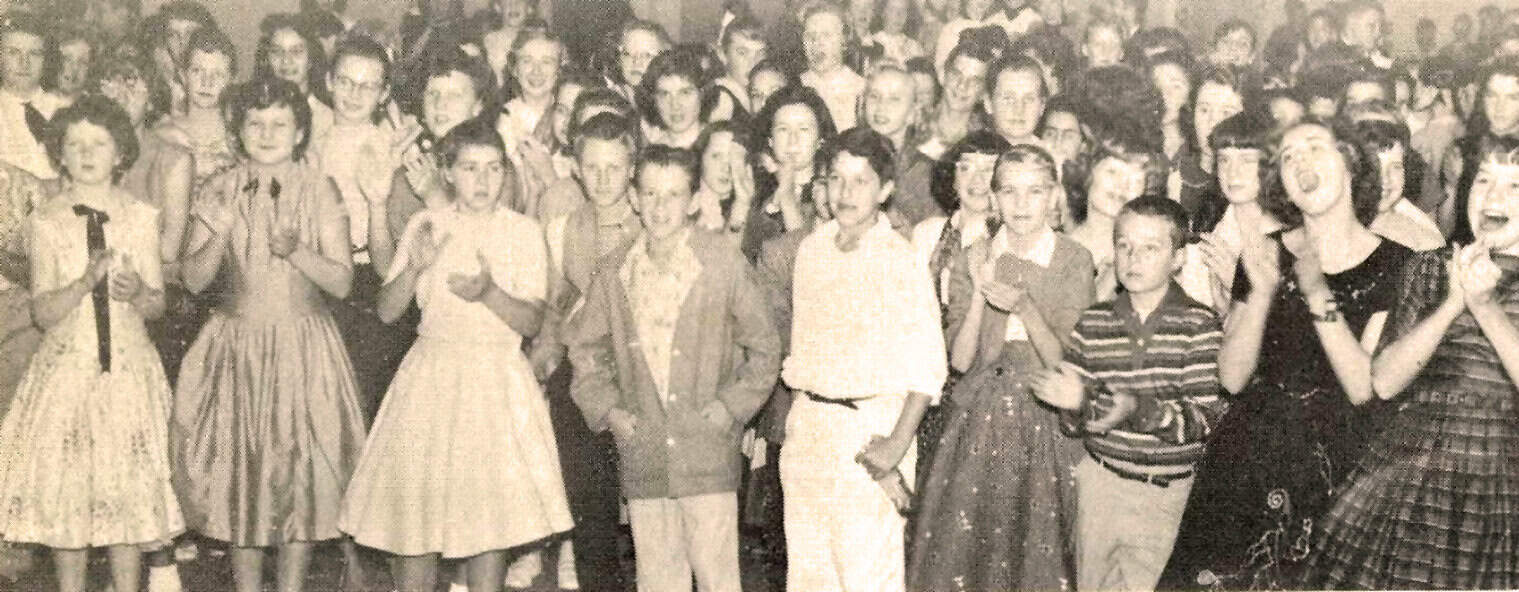 A lively group of children and teenagers at a school event, captured in black-and-white, showcasing vintage fashion and expressions of joy and excitement.
