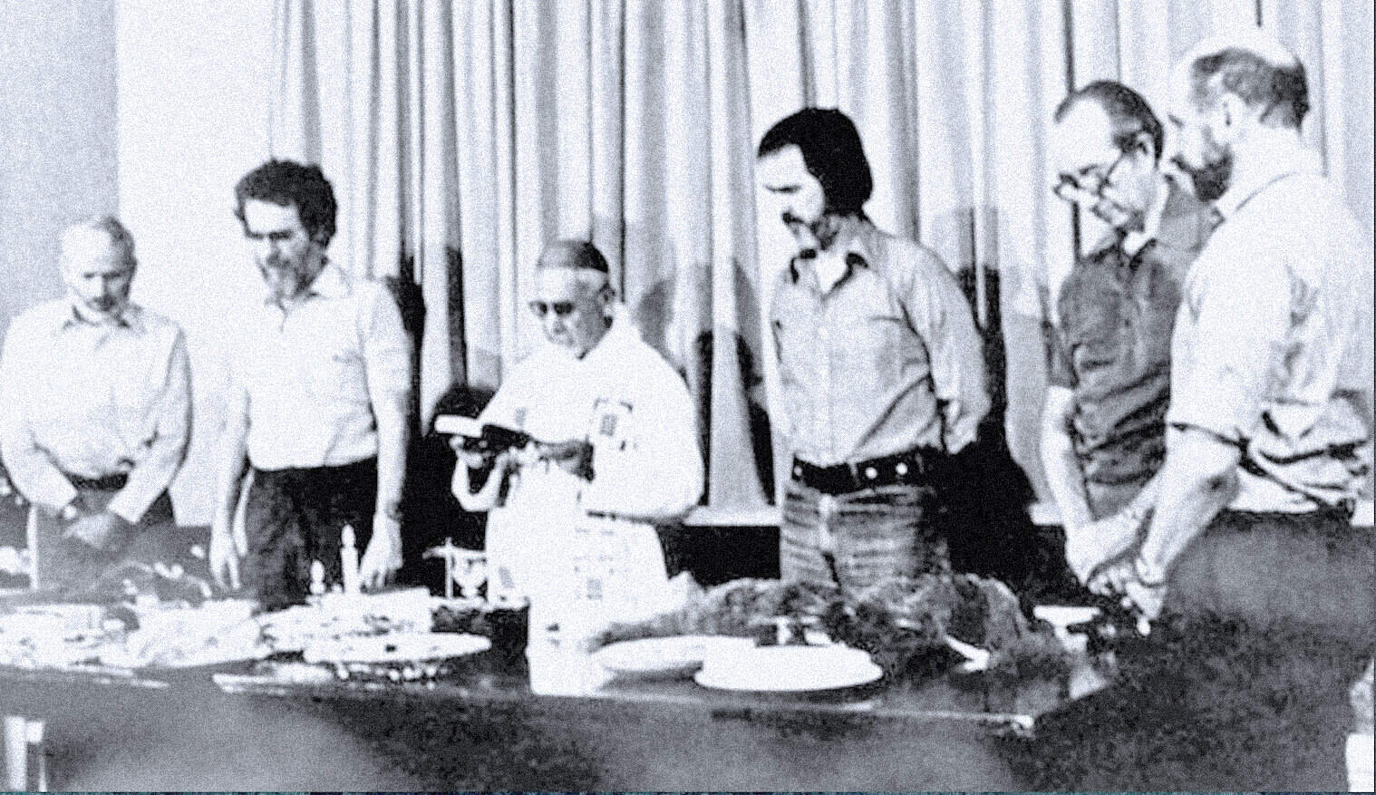 Vintage black and white photo of a group of men gathered around a table with food, celebrating or participating in a historical event related to news, music, or history.