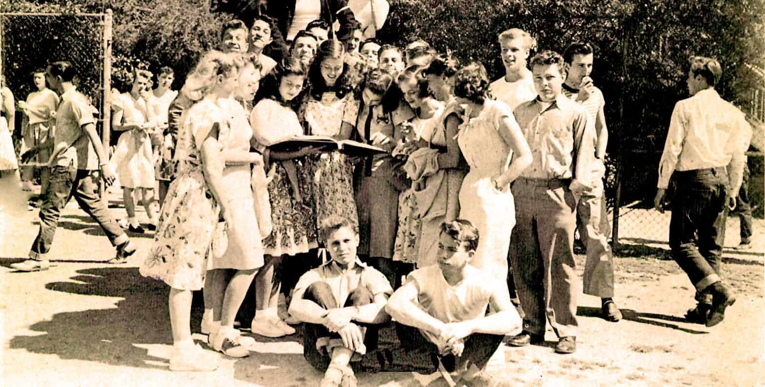 Vintage black and white photograph of school children gathered outdoors, reading and socializing, reflecting mid-20th century school life and community events.