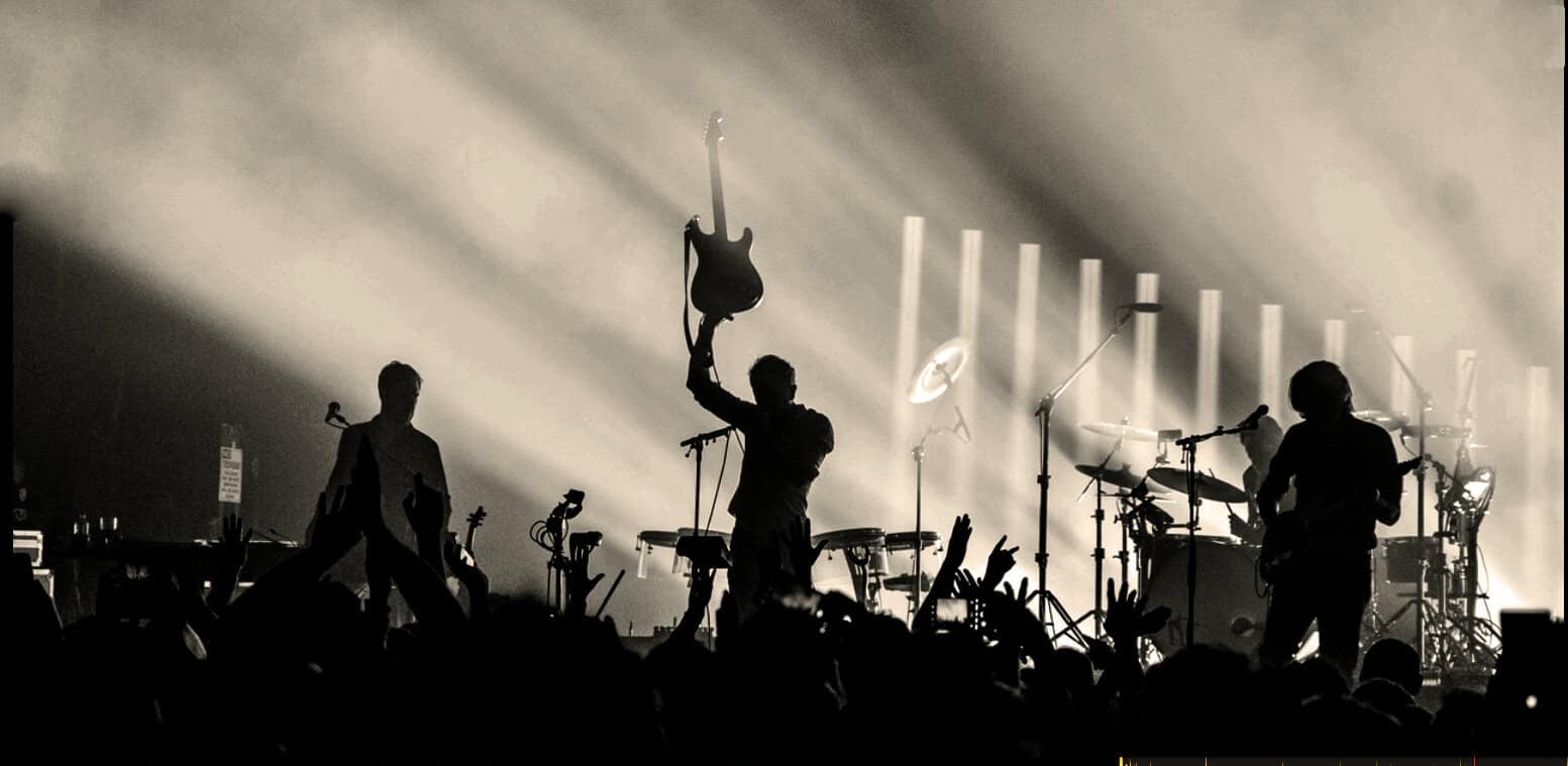 Energetic live music performance featuring a band on stage with striking lighting, guitars, drums, and enthusiastic audience at a concert or music event.