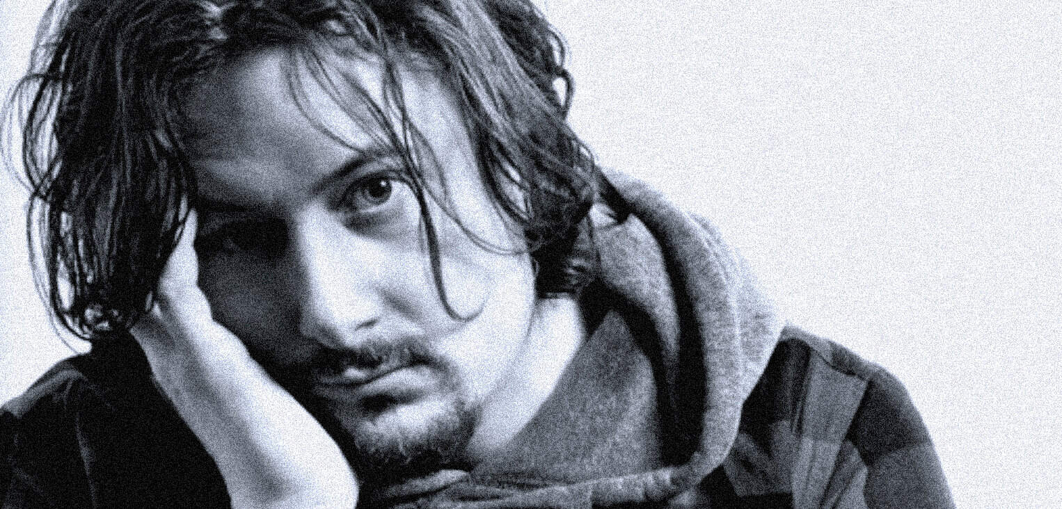 Portrait of a young man with wet hair, contemplative expression, wearing a hoodie and jacket, representing news, history, and music archive content.