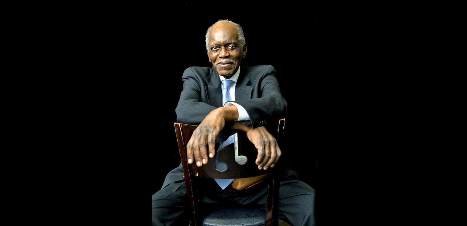 Elderly man in a suit posing with a music note cutout on a black background, representing historic news, music, and sound archive themes for Past Daily SEO content.