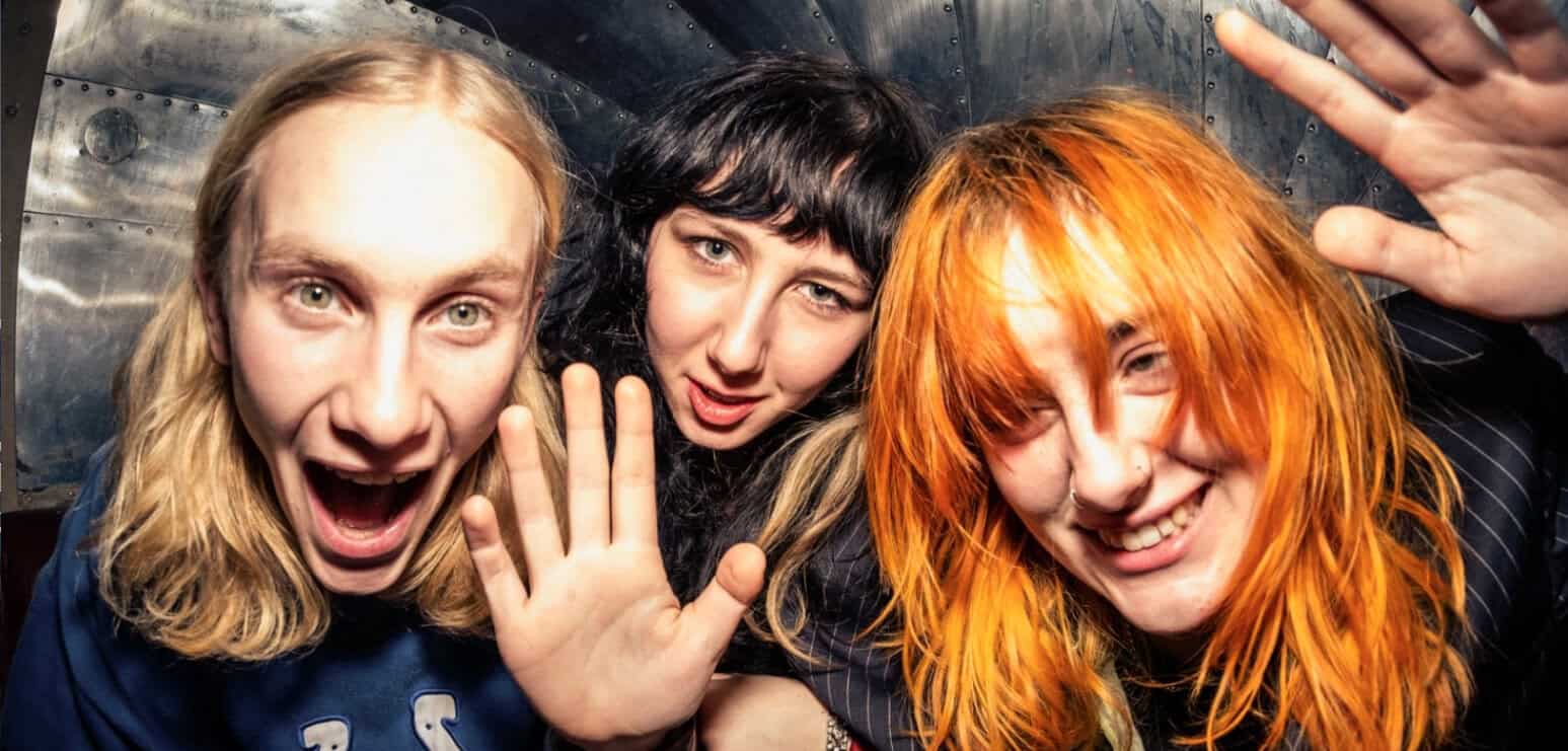 Vibrant photo of three excited young women with colorful hair, celebrating music and friendship, in a lively setting.
