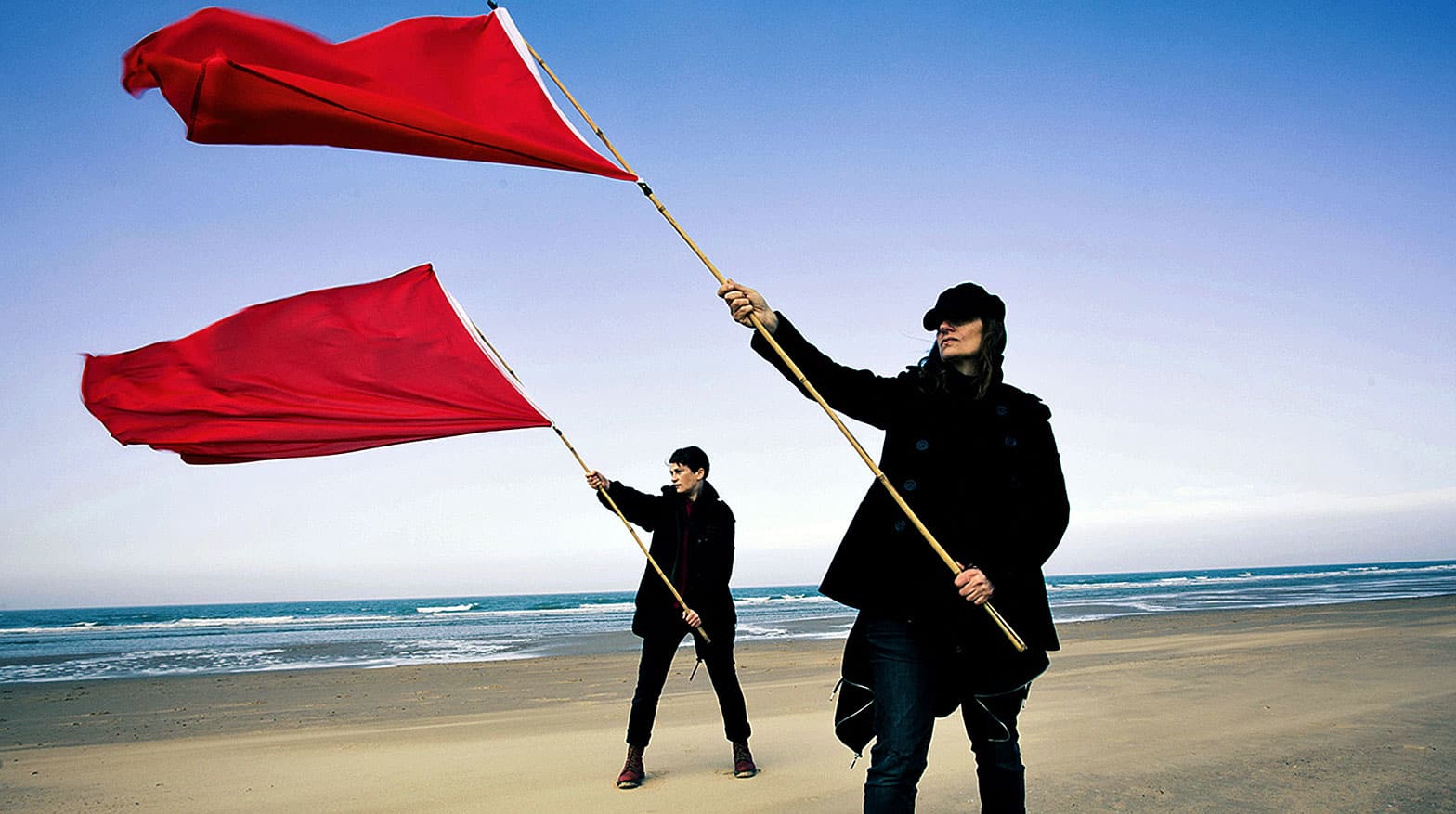Flags waving on beach with women holding red flags for protest and activism, scenic coastline, ocean waves, clear blue sky, social movement, freedom, and expression concepts, summer setting, outdoor event, empowerment, activism, public demonstration, protest scene, environmental awareness, community rally.