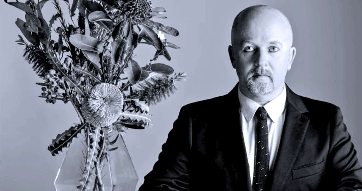 Vintage black and white photo of a man in a suit with a serious expression, next to a large floral arrangement, representing historical news, music archives, or media history themes.