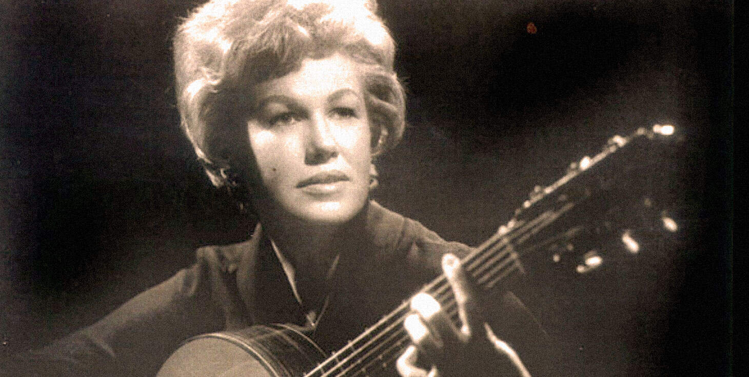 A black and white photo of a woman playing an acoustic guitar, capturing her in a musical performance related to classic music, news archives, and historic sound recordings.