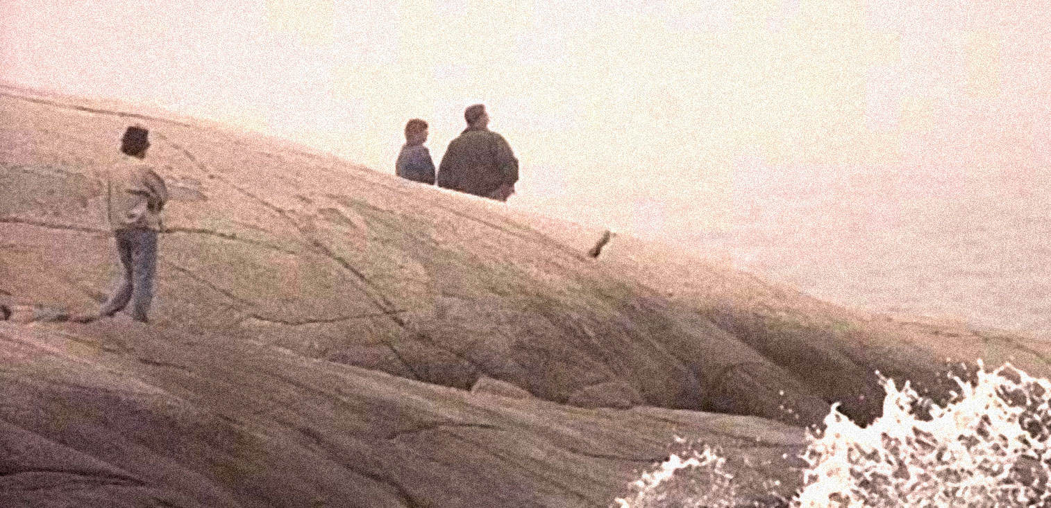 Photograph of people standing on rocky shoreline near water, vintage style, nostalgic, reflective scene with waves crashing, historical maritime setting, reminiscent of classic news and history archives, musical and cultural significance.