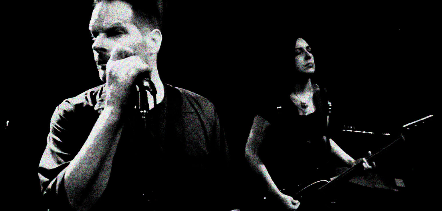 Male singer with microphone and female guitarist performing in a music archive setting.