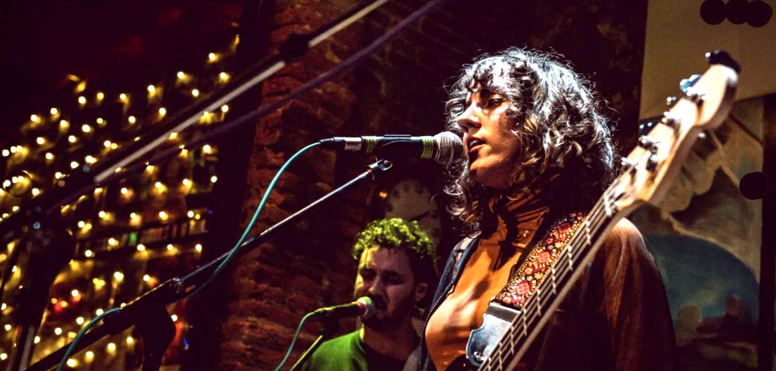 Live music performance featuring a female singer with a guitar at a cozy, intimate venue with warm lighting and decorative string lights.