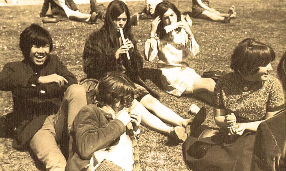 Young women relaxing on the grass in a vintage 1960s outdoor scene, enjoying music and conversation, capturing a nostalgic moment of social gatherings from the past.