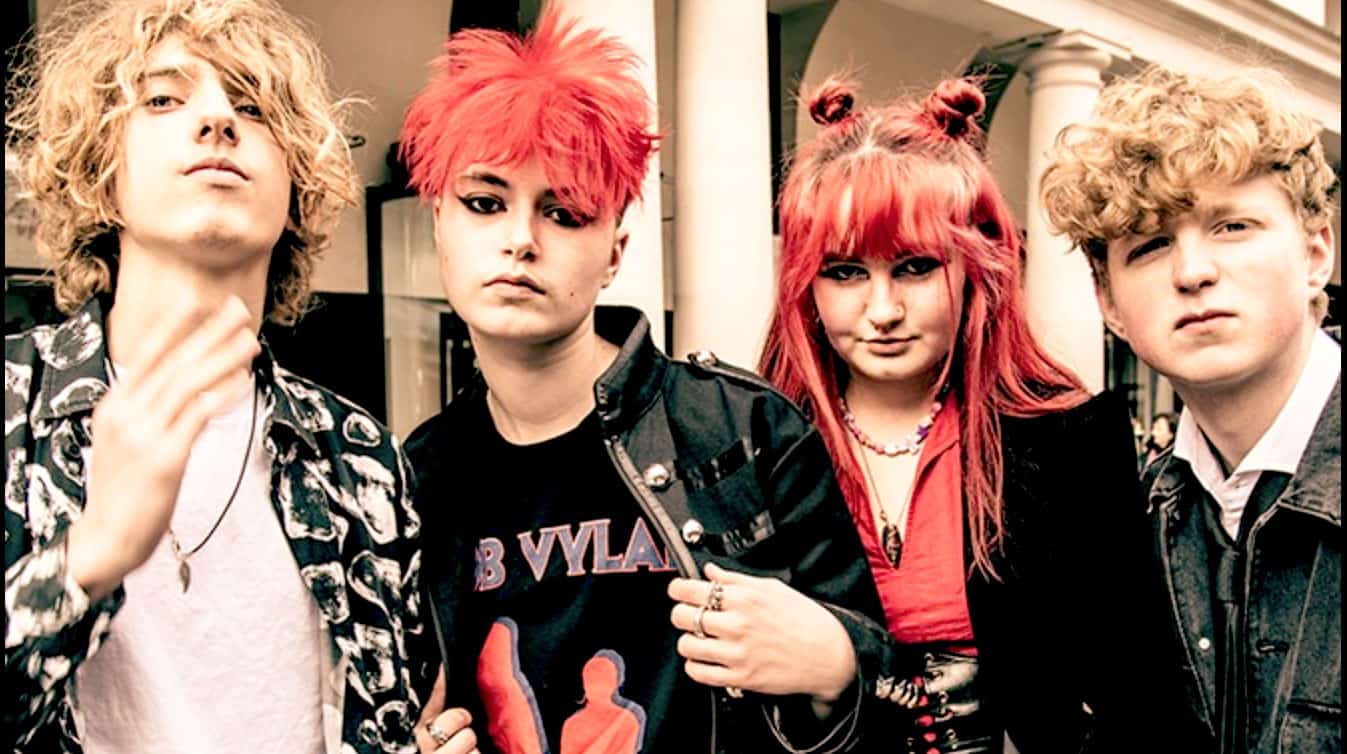 Punk rock band members with vibrant red and blonde hair, dressed in edgy clothing, standing outdoors during daytime, capturing alternative music culture.
