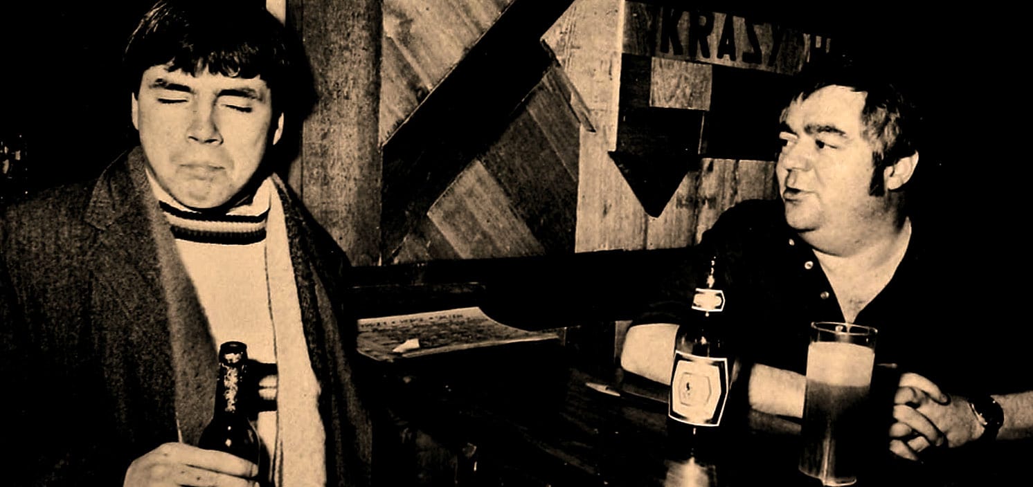 Vintage image of two men having a conversation in a bar, with bottles and glasses on the table, capturing a nostalgic music history scene, reflecting the audio archives of news, history, and music on Past Daily.
