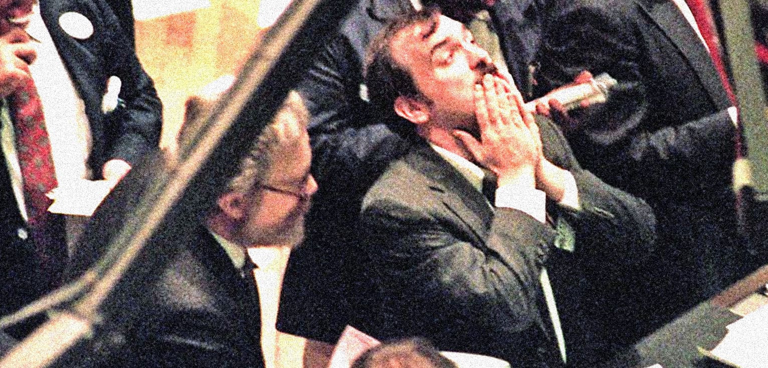 A man in a suit with glasses, passionately listening or reacting at a formal event or conference.