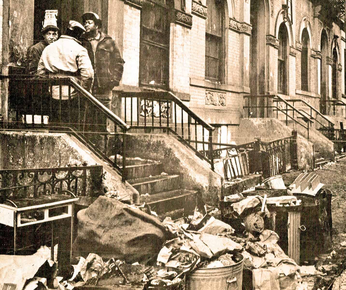 Disorderly street scene with trash and debris outside brownstone buildings, historic black-and-white photo, urban decay and community cleanup, vintage city life snapshot, relevance for news archives, history, and urban environment.